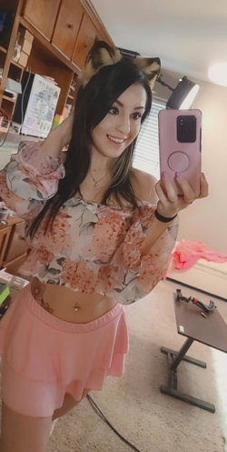 Brunette woman in floral crop top and pink skirt takes mirror selfie with cat ears, smiling in a room with shelves and