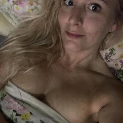 Blonde woman with long hair lying on a floral pillow, wearing a floral top that is pulled down, revealing bare breasts.