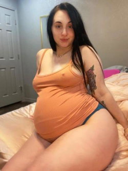 A curvy woman with long dark hair sits on a bed wearing an orange tank top and blue underwear, showing visible tattoos