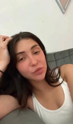 Brunette woman with a curvy build lying on a gray couch, wearing a white tank top and resting her head on her hand.