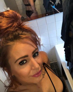 Redheaded woman with a nose ring and shoulder tattoo takes a selfie in a bathroom mirror, wearing a black top with thin