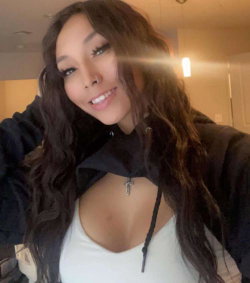 A woman with long dark wavy hair smiles in a selfie, wearing a white top under a black hoodie in a warmly lit indoor