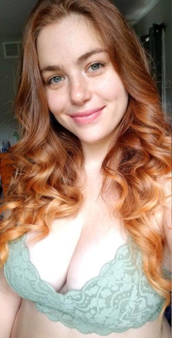 A woman with long wavy red hair and green eyes smiles softly while wearing a light green lace bra, captured in a
