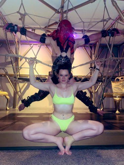 A woman in a bright yellow bikini sits cross-legged on the floor while another person in red and black gear stands