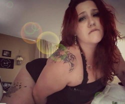 A woman with red hair and visible tattoos on her shoulder and arm wears a black top, posing indoors with soft lighting
