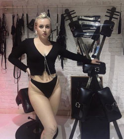 Blonde woman in black crop top and thong posing in a room with leather gear and bondage equipment.