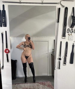Blonde woman in black lingerie and thigh-high boots takes mirror selfie in a room with BDSM gear on the walls.
