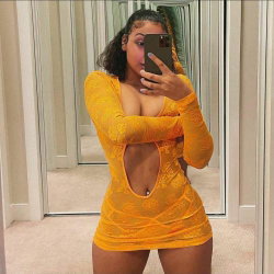 A curvy woman in a yellow lace dress with a back cutout takes a mirror selfie in a carpeted room.