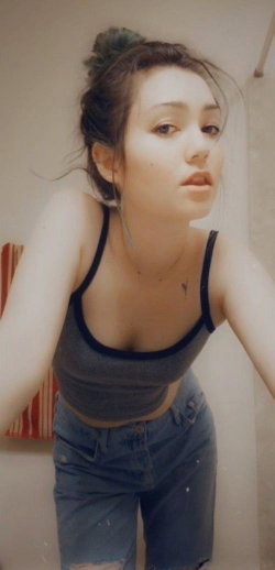 A young woman with dark hair in a messy bun wears a gray crop top and denim shorts, leaning forward in a casual indoor