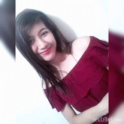 A woman with long dark hair smiles in a red off-shoulder top, leaning against a white wall in a close-up selfie.