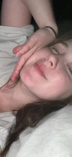 A woman with long dark hair lies on her side, resting her head on a white surface, hand near her face, wearing