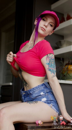 A woman with purple hair and tattoos wears a pink crop top and denim shorts, posing indoors with one hand lifting her