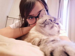 Woman with glasses and a cat lying on a bed in a well-lit room.