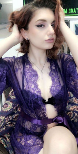 Brunette woman in a sheer purple lace robe with black lingerie underneath, posing indoors with hands in her hair.