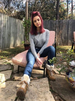 A woman with purple hair sits on a patio chair wearing a gray sweater, blue jeans, and fur-lined boots in a garden