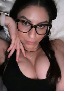 Brunette woman with glasses wearing a black top, lying on a white surface, hand near her face in a close-up selfie.