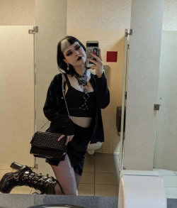 A woman with dark hair and bangs takes a mirror selfie in a bathroom, wearing black goth-style clothing, thigh-high