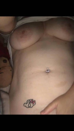 Close-up of a person's torso showing bare breasts, a belly button piercing, and a small tattoo on the lower abdomen,