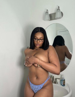 A curvy woman with dark hair and glasses stands in a bathroom, wearing blue patterned thong underwear and covering her