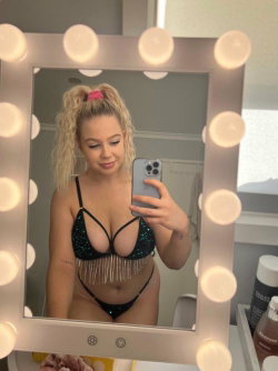 Blonde woman in a sequined black bra and thong takes a mirror selfie with a phone, framed by vanity lights.
