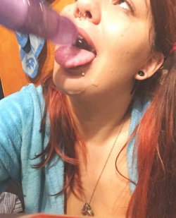 Redheaded woman with piercings and a blue robe takes a selfie while holding a purple toy to her mouth.