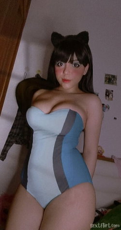 A woman with dark hair and cat ears wears a blue and gray strapless bodysuit, posing in a bedroom setting.
