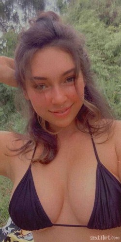 A woman with dark hair in a bun wears a black bikini top, smiling at the camera outdoors in a grassy area.