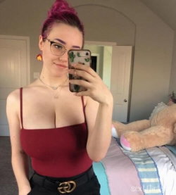 A woman with magenta hair in a bun and glasses takes a mirror selfie in a red tank top and black pants, standing in a