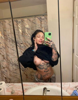 A woman with dark hair and multiple tattoos takes a mirror selfie in a bathroom, wearing a black top pulled down to