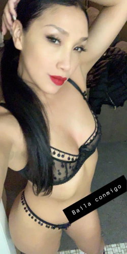 A woman with long dark hair and red lipstick poses in black lace lingerie, holding her hair back with one arm raised.