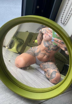 A tattooed person with a curvy build kneels in front of a round mirror, wearing a gray thong and holding a phone,