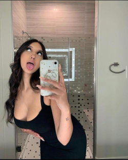 Brunette woman in black top takes mirror selfie in bathroom, sticking out tongue with hand on hip.
