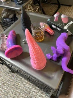 Various colorful sex toys and a glass of amber liquid on a metal table in a casual indoor setting.