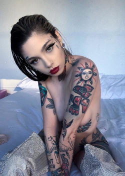 A woman with dark hair and bold makeup sits on a bed, her upper body bare and covered in colorful tattoos, looking