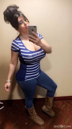 Curvy woman in a blue and white striped top, jeans, and boots poses for a mirror selfie in a room with patterned carpet.