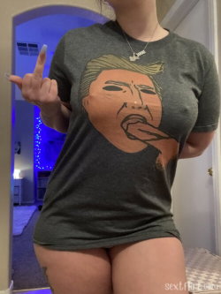 Curvy woman in a graphic t-shirt posing with one hand raised, showing thong underwear and tattoo on thigh, indoors with