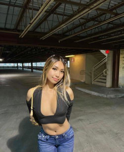 A woman with long blonde-highlighted hair wears a black cutout top and blue jeans, standing in an industrial parking