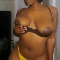 Curvy woman with dark skin wearing a brown bra and yellow underwear, standing indoors with one hand near her chest.
