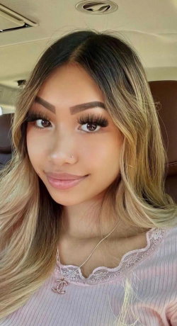 A woman with long ombre hair and makeup smiles in a car selfie, wearing a pink lace-trimmed top.