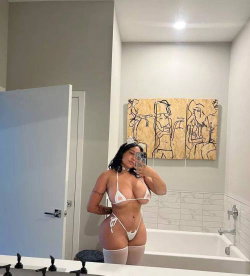 Curvy woman in white bikini and thigh-high stockings takes mirror selfie in modern bathroom with artwork on wall.