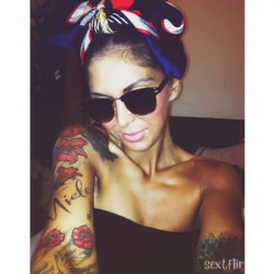 A woman with dark hair wearing a colorful headscarf and sunglasses, showing tattooed arms in a black top indoors.