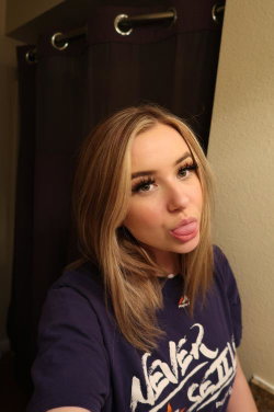 Blonde woman with shoulder-length hair making a playful face in a purple t-shirt, taking a selfie indoors near a dark