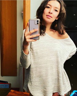 Woman with shoulder-length dark hair takes a mirror selfie in a loose off-the-shoulder sweater, indoors.