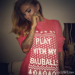 A woman with long ombre hair and visible arm tattoos wears a red sweater with festive text, posing indoors with hand