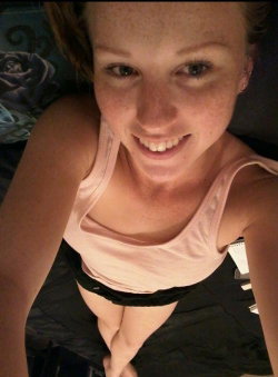 A smiling woman with fair skin and freckles takes a close-up selfie, wearing a light pink tank top and black bottoms,