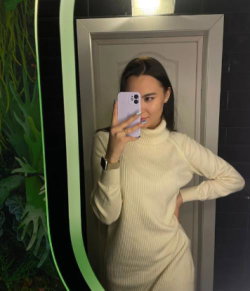 Woman in a cream ribbed turtleneck dress poses in a mirror selfie, hand on hip, dark hair pulled back, in a room with