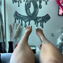 A person with tattooed legs and painted toenails sits on a couch, feet raised toward a wall with a dripping Chanel logo