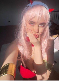 A person with pink hair and devil horns poses in a red top and gold arm bands, taking a selfie indoors.