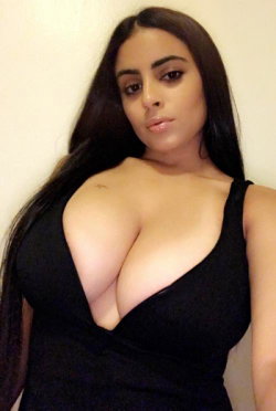 Brunette woman with long dark hair wearing a black plunging top, posing indoors against a plain wall.