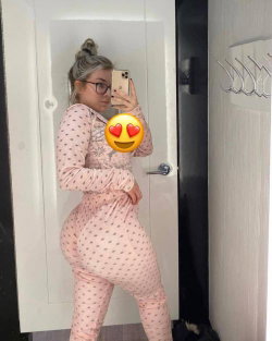 A woman with light hair in a bun and glasses takes a mirror selfie wearing pink patterned loungewear, posing with one